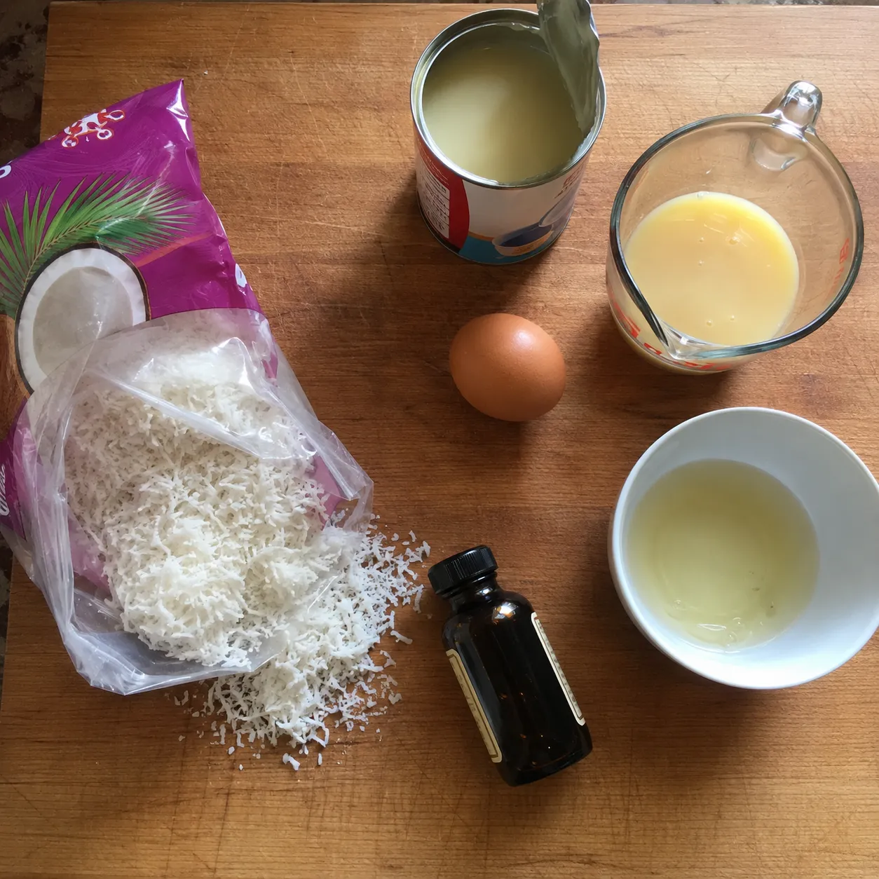 Sweetened shredded coconut, condensed milk, an egg, egg white and vanilla laid out on a wooden board