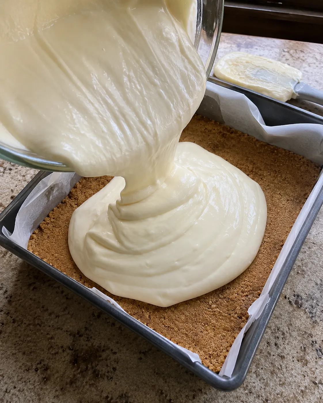 Smooth cheesecake filling being poured from a glass bowl over a baked graham crust in a parchment-lined square pan