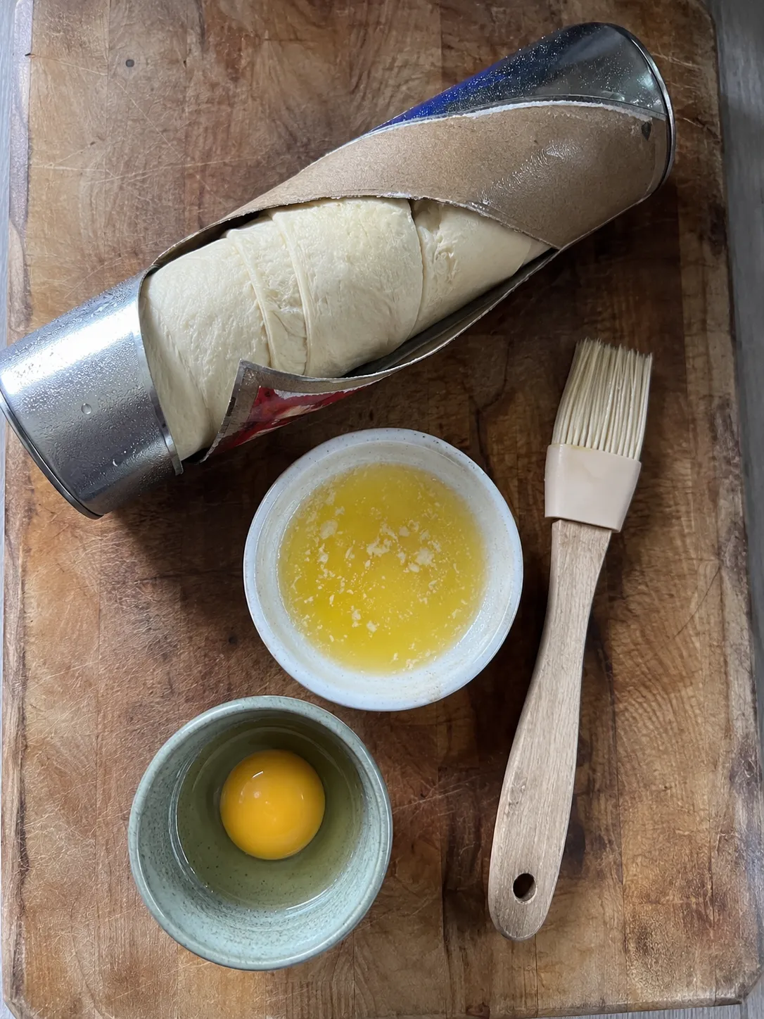 An opened tube of refrigerated crescent roll dough on a wooden board beside melted butter, an egg yolk and a pastry brush