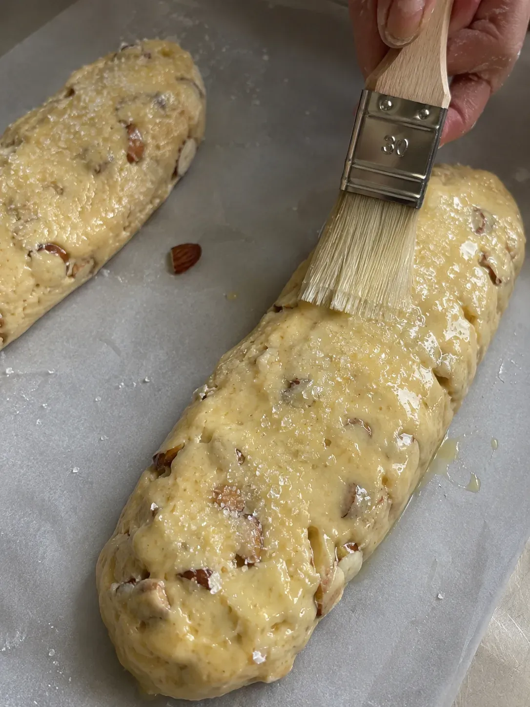 Brushing egg wash onto a shaped almond biscotti log and sprinkling it with coarse sugar