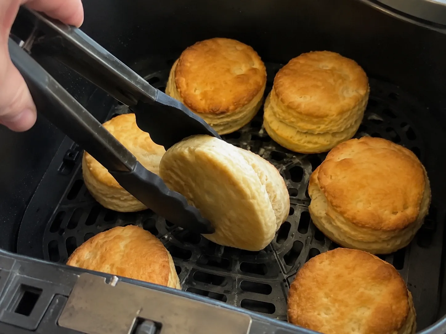 Tongs turning a golden-topped biscuit over in an air fryer basket, its pale underside showing