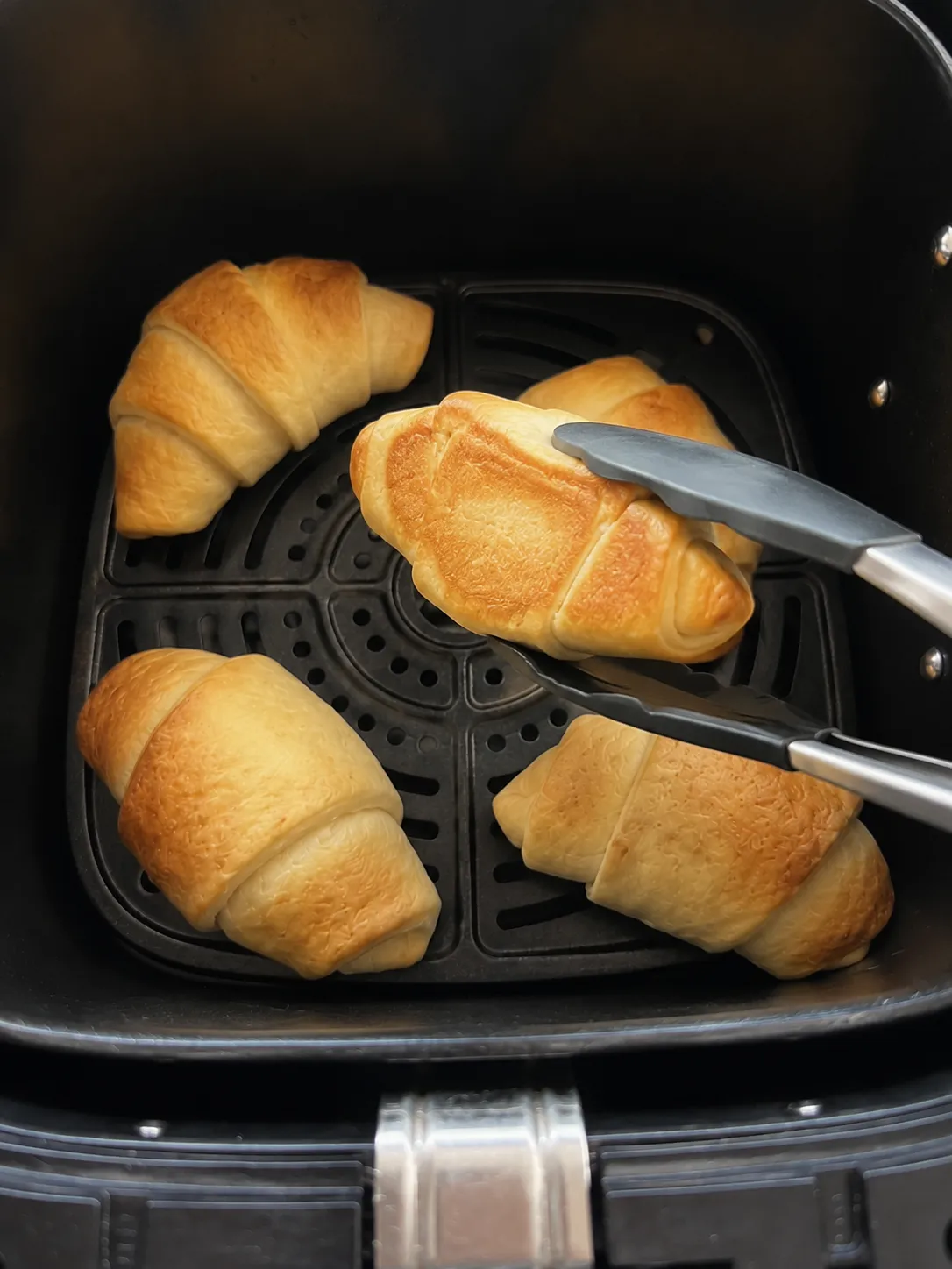 Tongs lifting one baked crescent roll in the air fryer basket to check that the underside has browned