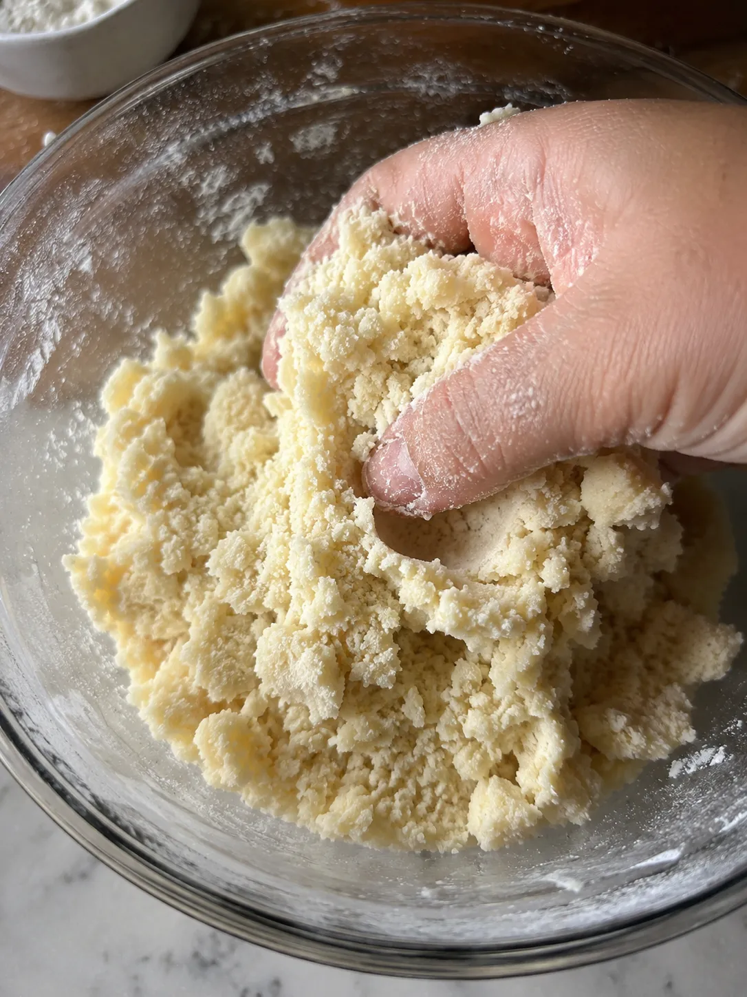 A hand squeezing crumbly shortbread dough in a glass bowl to show it holding together