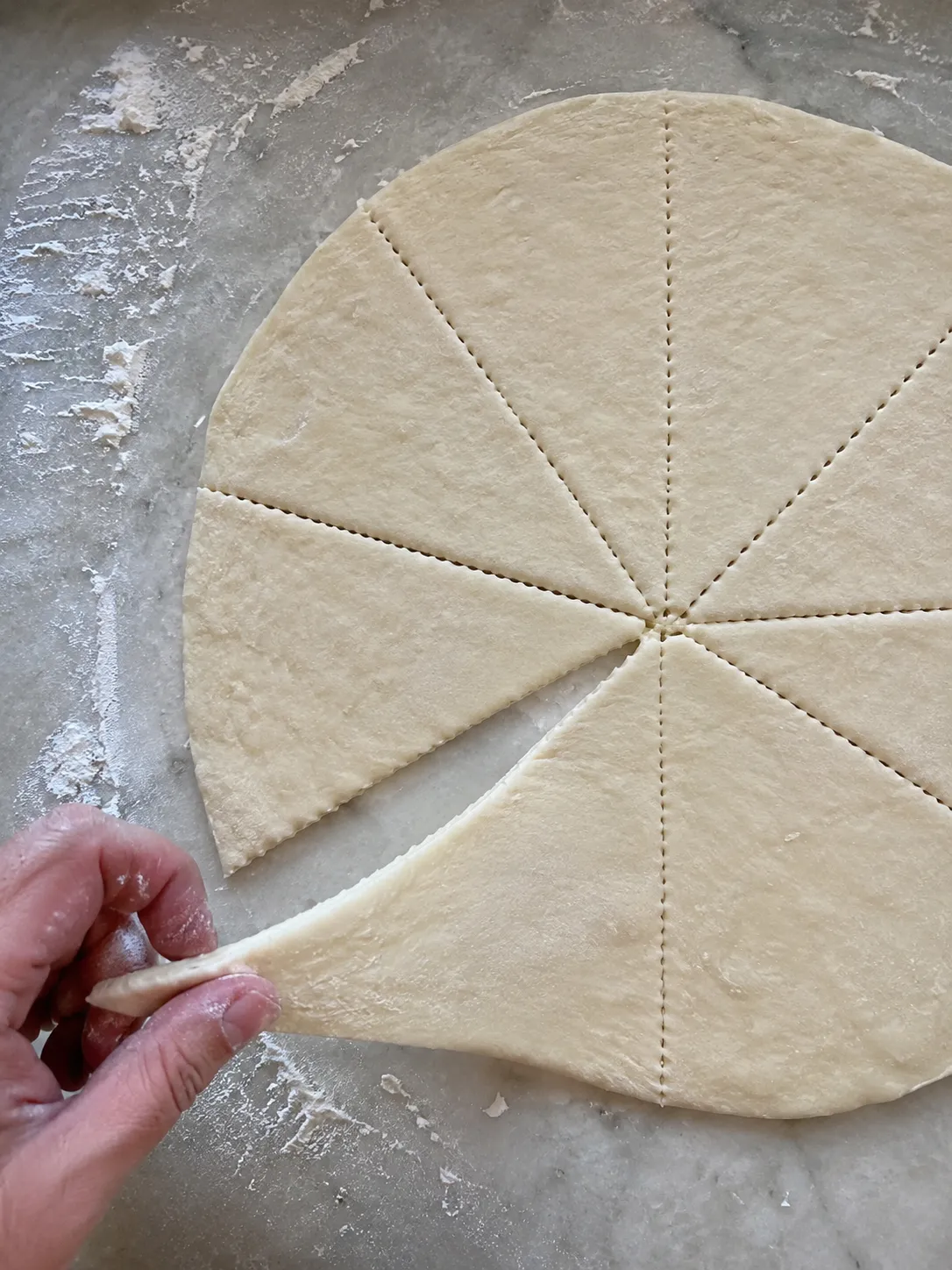 A hand peeling one triangular wedge away from a round of perforated crescent roll dough on a floured counter