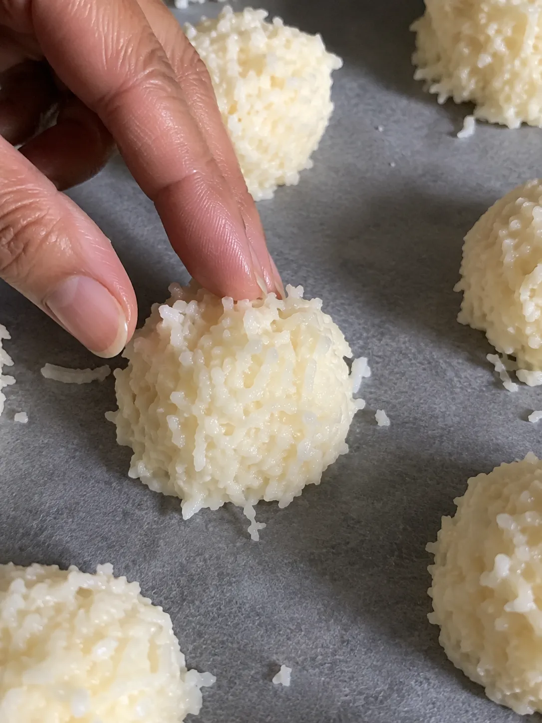 Damp fingers pressing a stray coconut shred back into a scooped macaroon mound on parchment