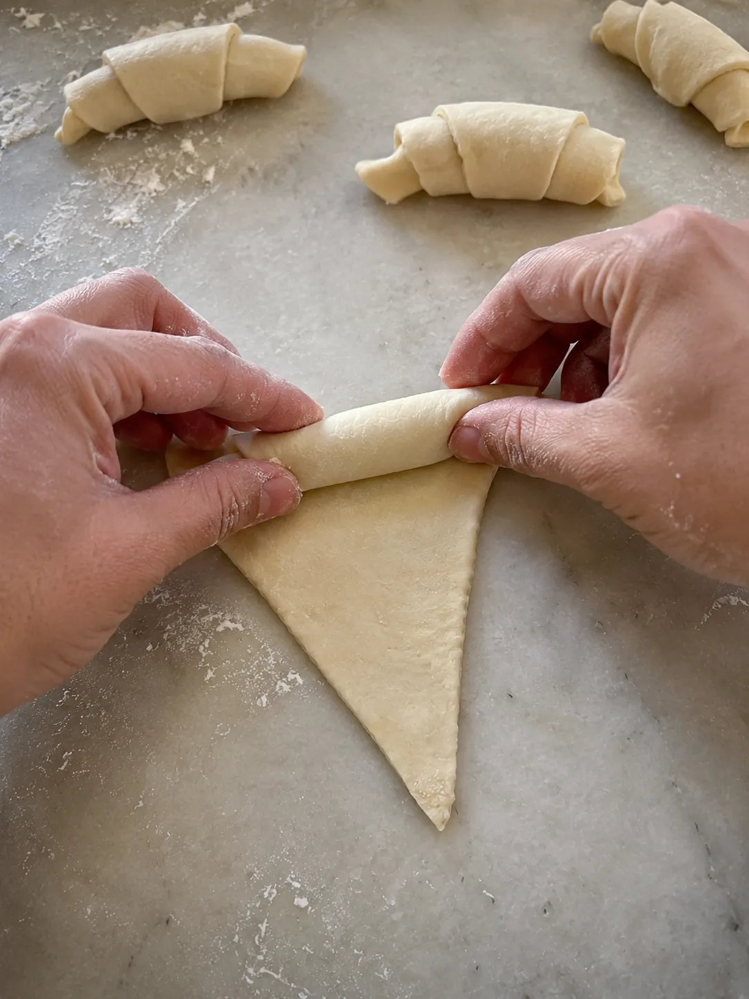 Two hands rolling a triangle of crescent dough loosely from the wide end, with finished rolls resting behind