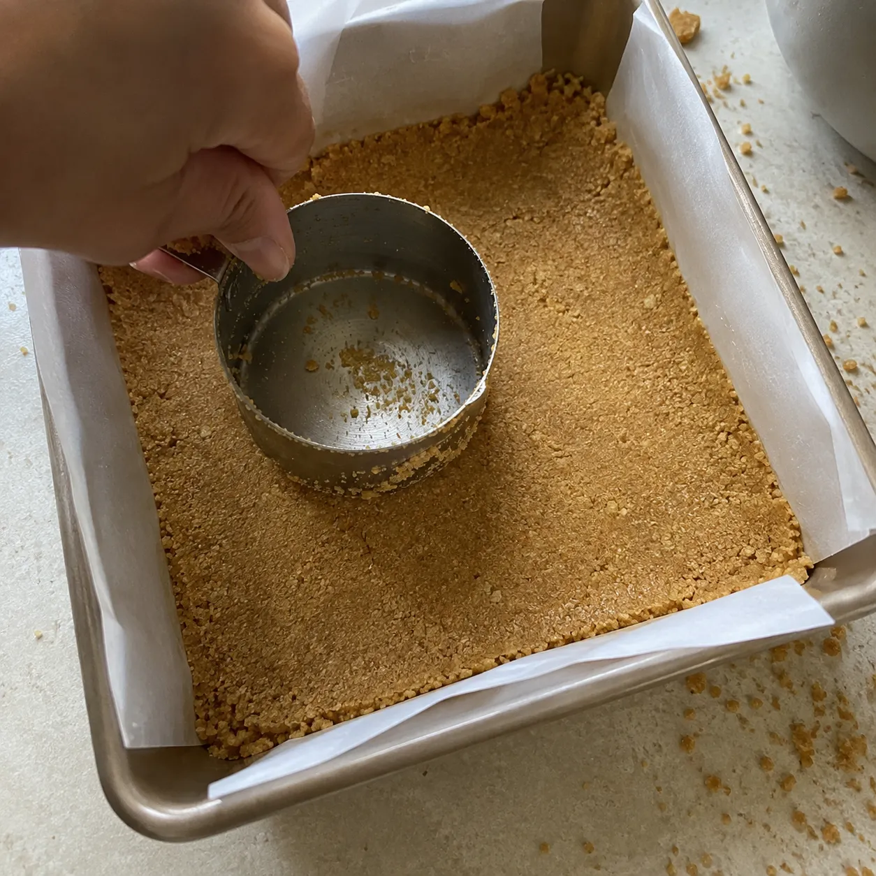 A hand pressing graham cracker crumbs into an even crust using the flat base of a metal measuring cup in a parchment-lined square pan