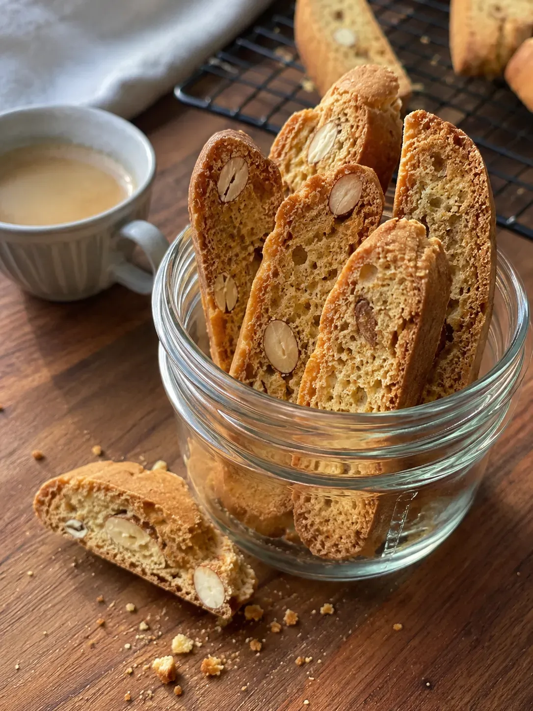 Almond biscotti standing in a glass jar beside a cup of coffee, with one broken slice on the table