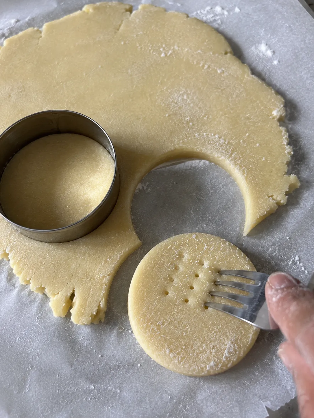 Rolled shortbread dough with a round cutter and a cut disc being pricked with a fork