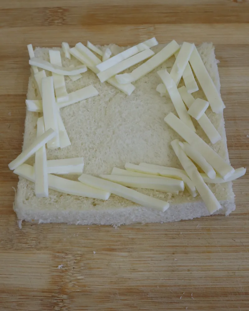 Sticks of cheese arranged around the border of a slice of white bread on a wooden board