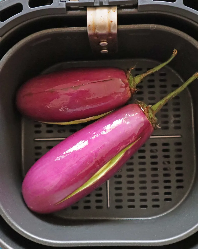 Two oiled eggplants slit lengthwise sitting in an air fryer basket before roasting