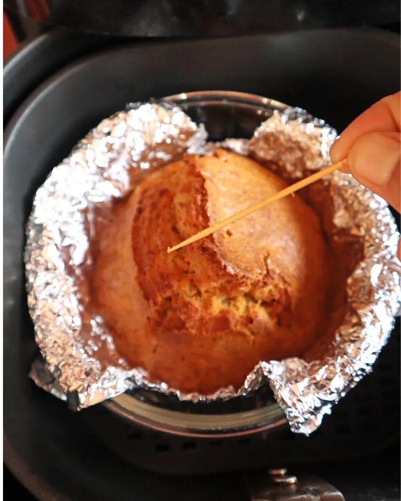 Baked carrot cake in a foil-lined pan inside an air fryer basket being tested with a toothpick