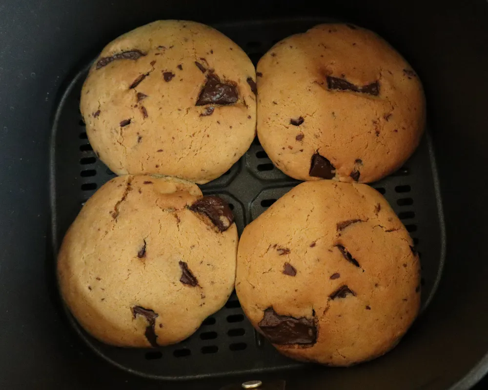 Four chocolate chip cookies baked in an air fryer basket, spaced apart on the bare perforated floor
