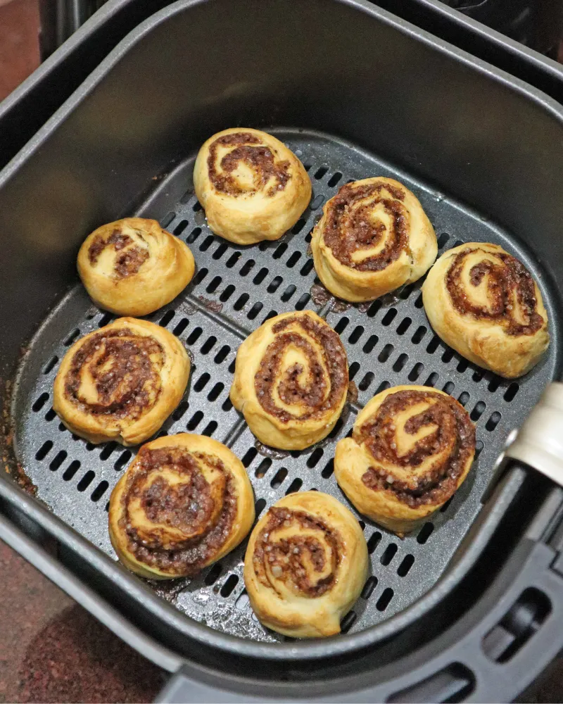 Nine baked cinnamon rolls with golden swirls in an air fryer basket