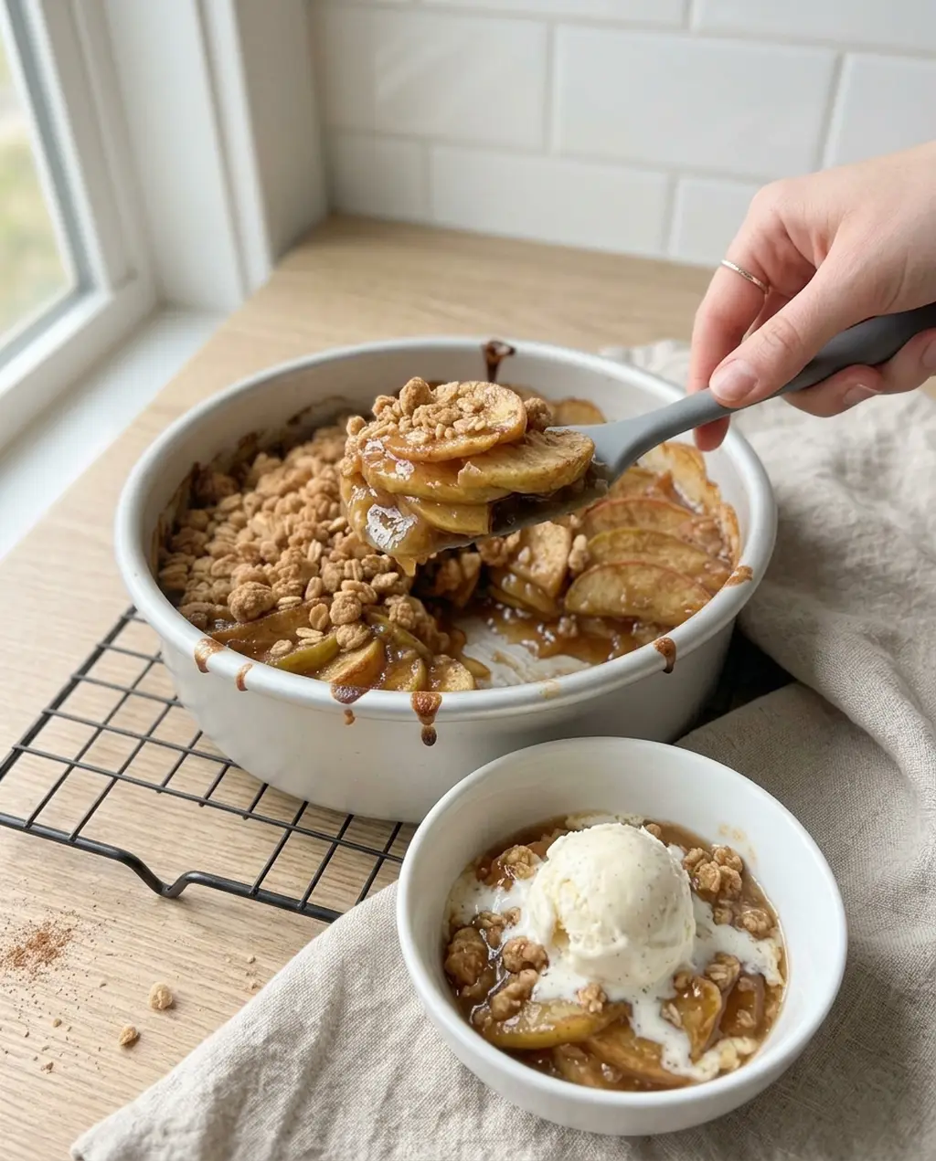 A spoon lifting a scoop of air fryer apple crisp with jammy apple slices and oat topping from a white dish, beside a bowl topped with vanilla ice cream