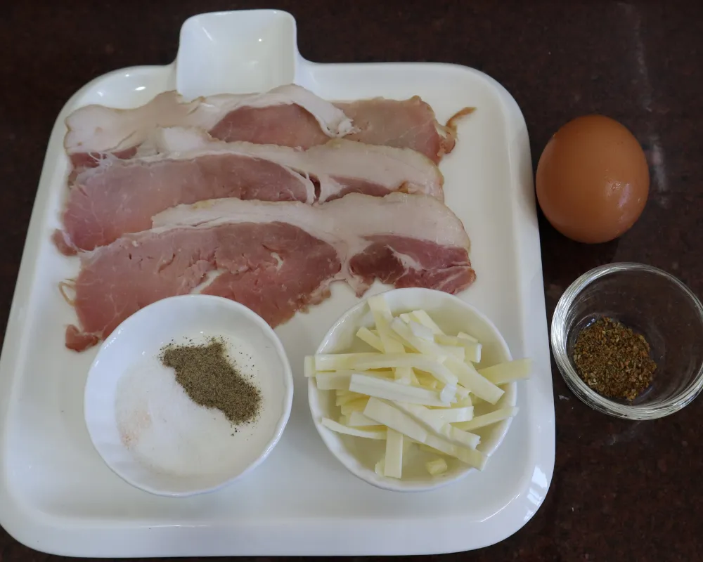Bacon rashers, an egg, salt and pepper, cheese sticks and dried herbs laid out on a white tray
