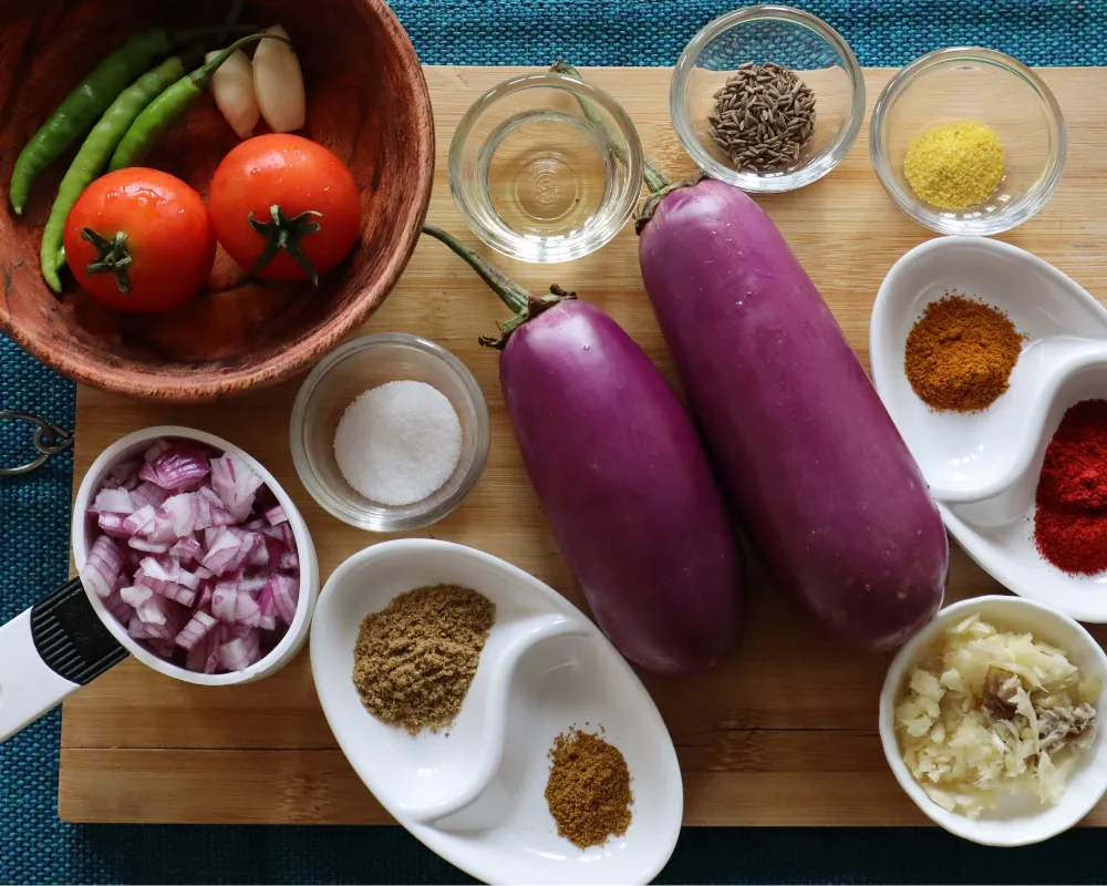 Baingan bharta ingredients on a board: two eggplants, tomatoes, green chilies, garlic, red onion, ginger and ground spices