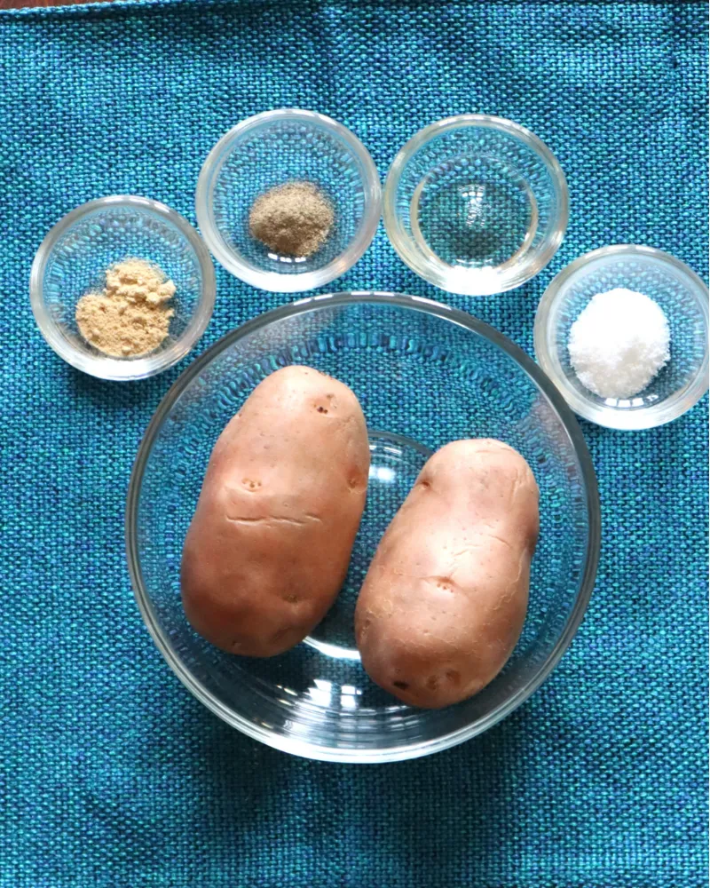 Two russet potatoes in a glass bowl with small bowls of oil, salt, pepper and garlic powder