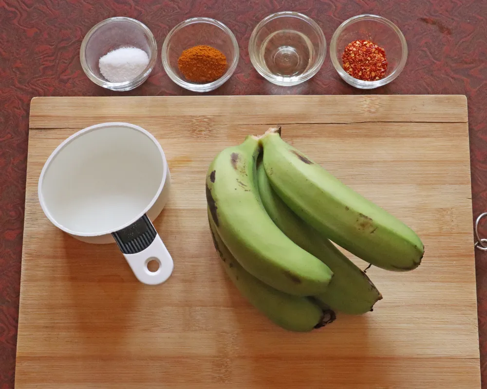 Green bananas on a board with bowls of salt, turmeric, oil and chili flakes and a cup of water