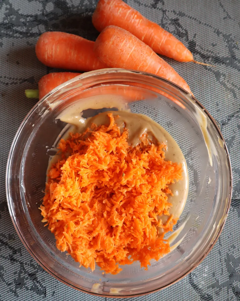 Grated carrot piled on top of wet cake batter in a glass bowl before folding