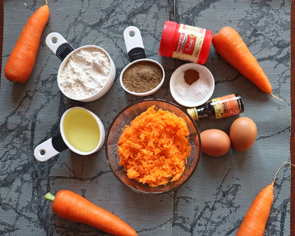 Carrot cake ingredients laid out: grated carrot, flour, sugar, oil, eggs, cinnamon and vanilla