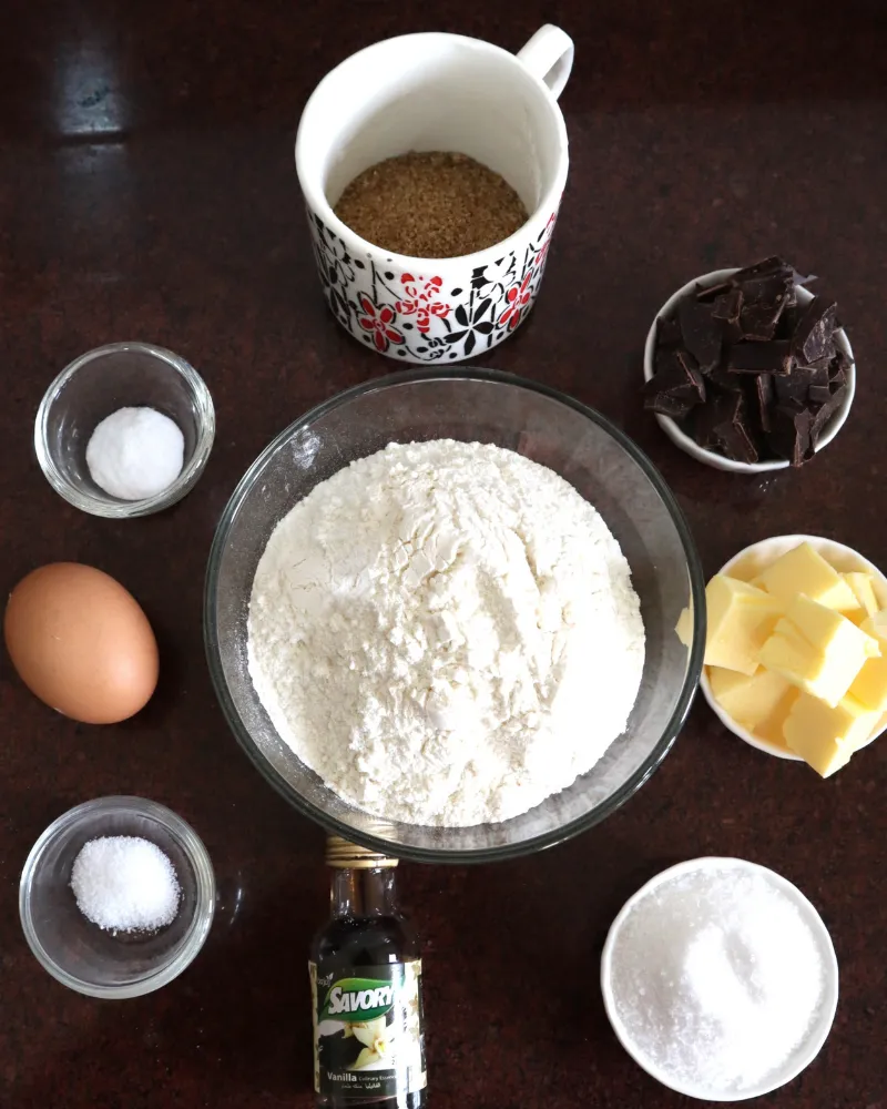 Chocolate chip cookie ingredients measured into bowls: flour, brown sugar, chopped chocolate, butter, egg, vanilla, salt and baking soda