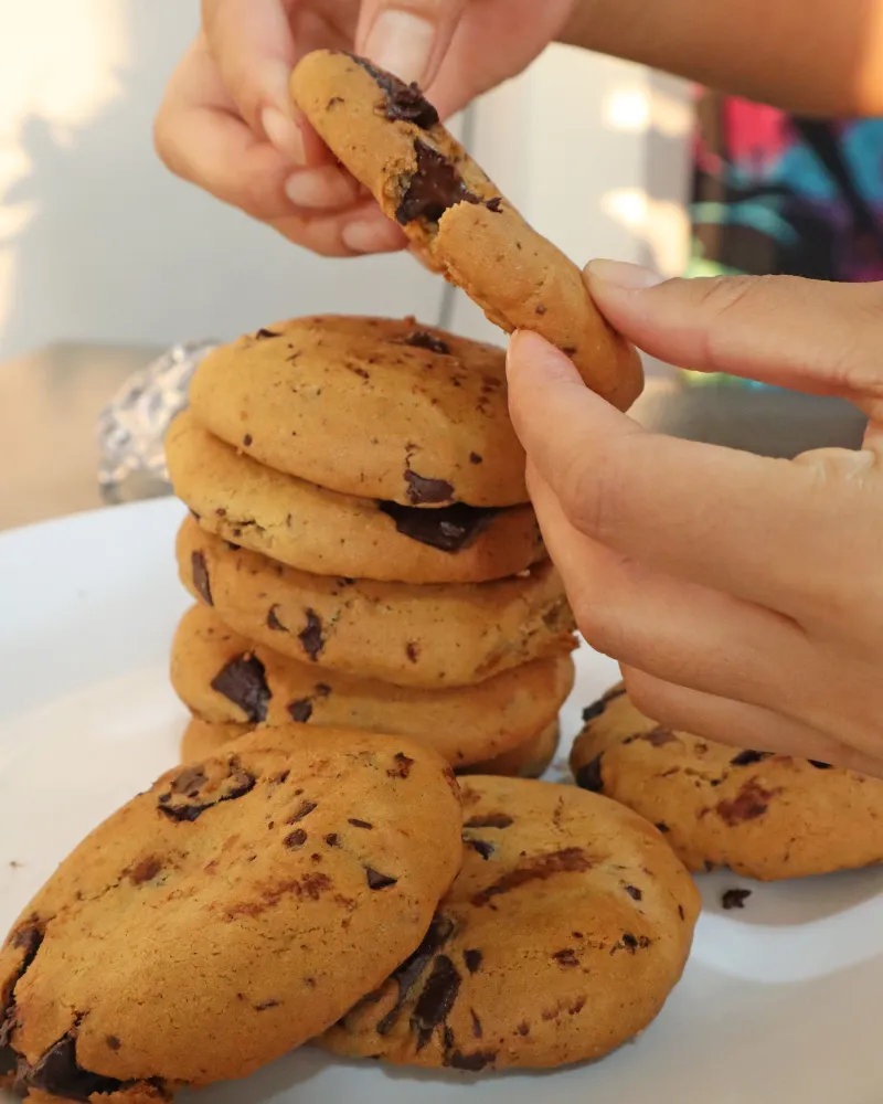 Hands pulling apart an air fryer chocolate chip cookie above a stack of cookies on a white plate