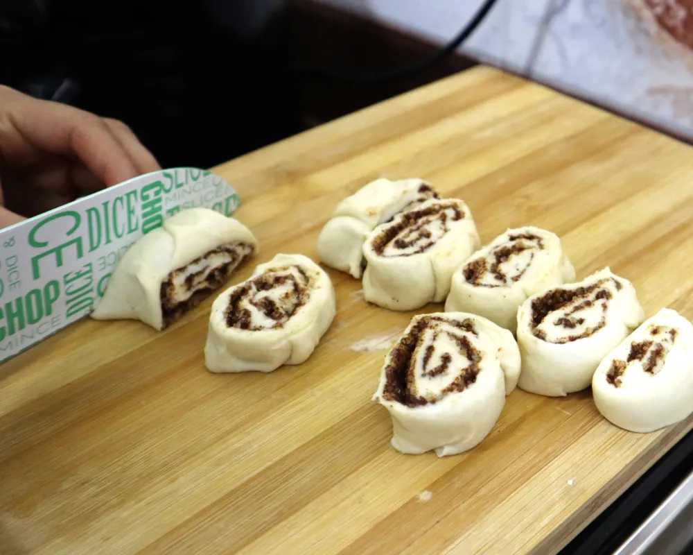 A rolled log of cinnamon roll dough being sliced into individual rolls on a wooden board