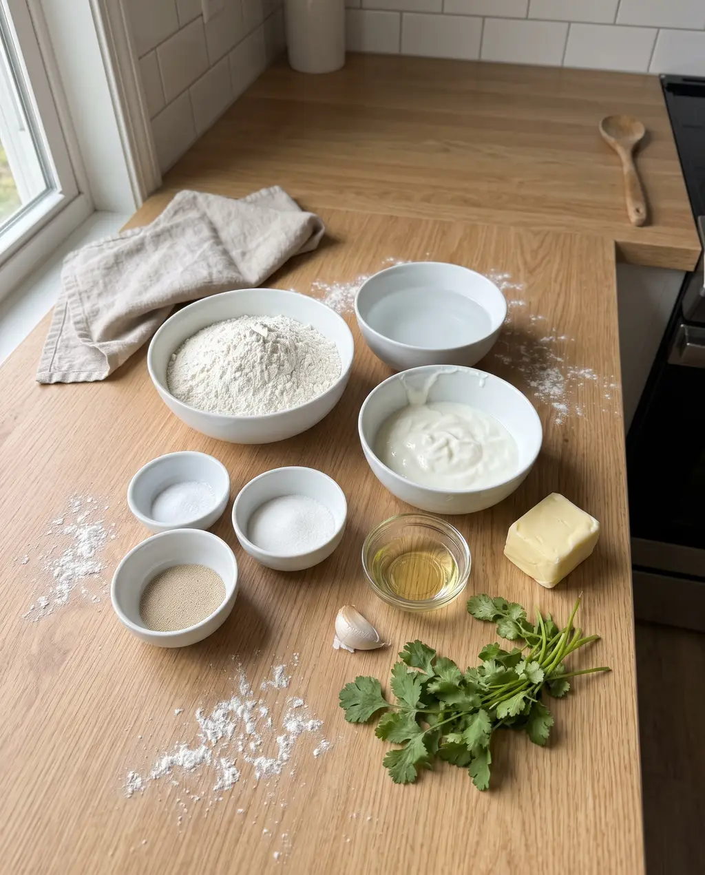 Naan ingredients on a wooden counter: flour, yogurt, warm water, yeast, sugar, salt, oil, butter, a garlic clove and fresh cilantro