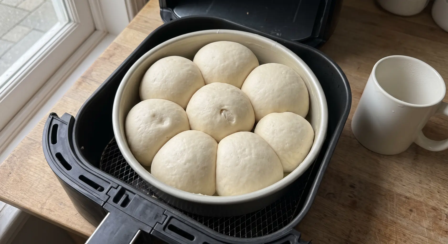 A pan of risen dinner rolls fused together inside an air fryer basket with a mug of hot water beside it