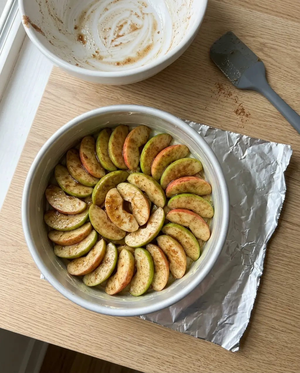 Seasoned apple slices shingled in a round white baking dish next to a sheet of foil