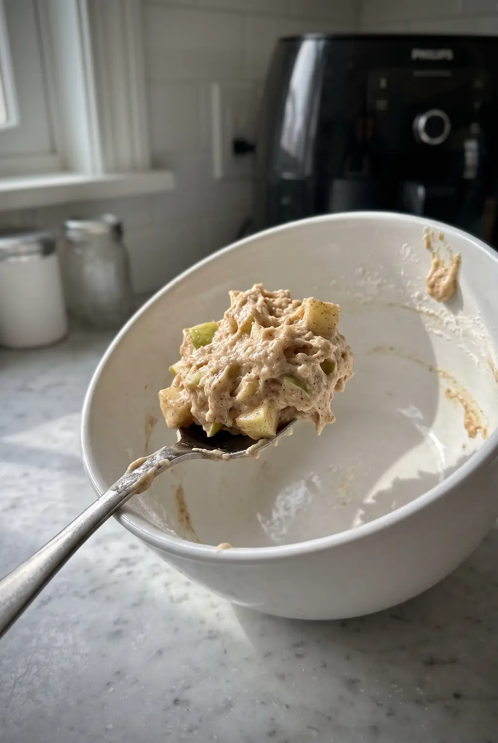 A spoonful of thick apple-studded fritter batter held on its side above the mixing bowl, holding a shaggy mound without dripping