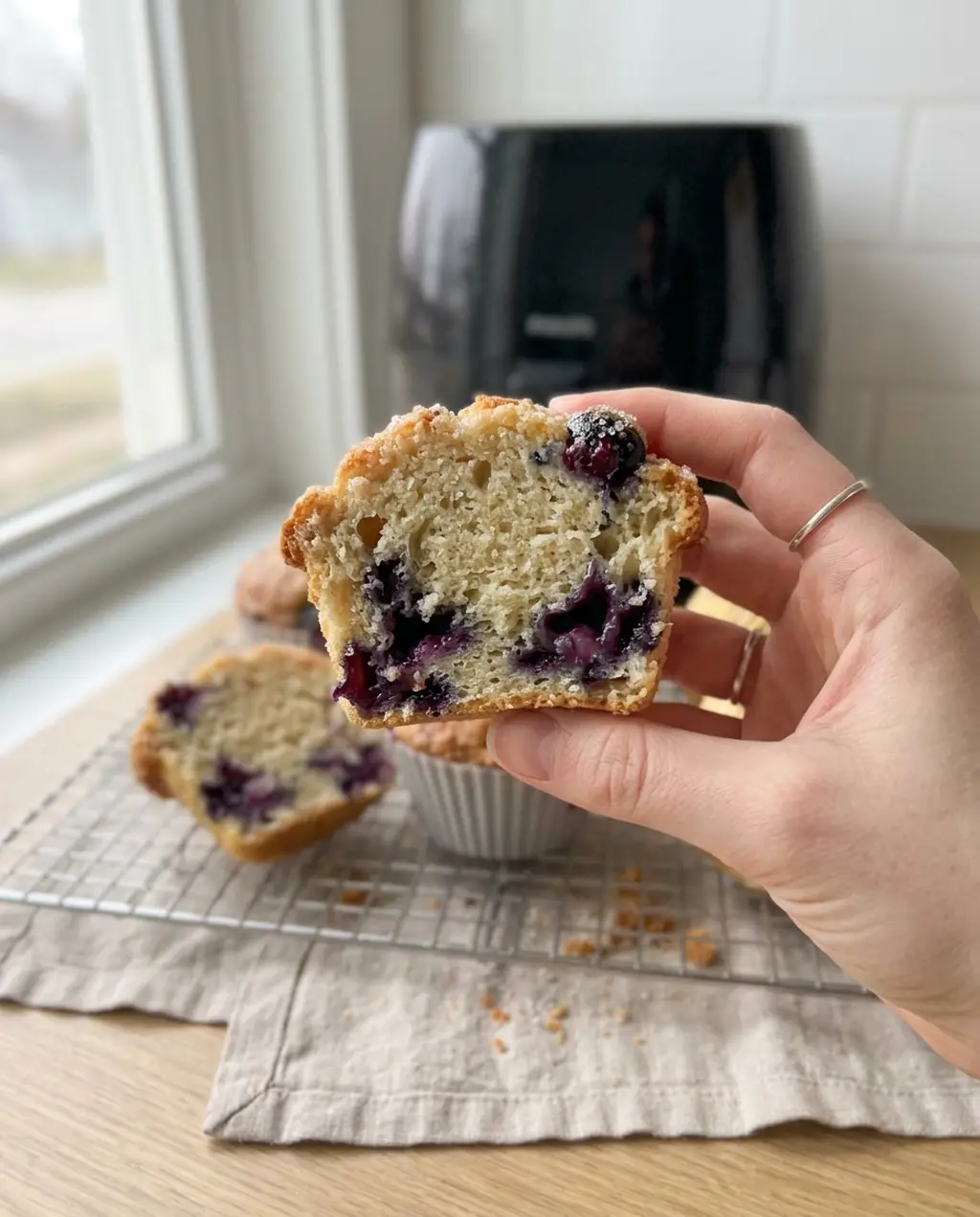 A hand holding a boxed mix blueberry muffin split open to show a tender crumb with blueberries spread through it