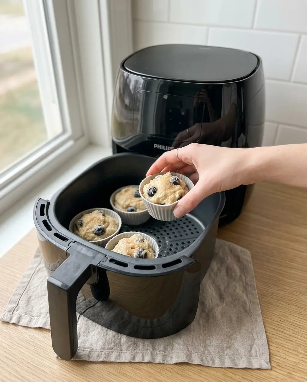 A hand placing a silicone cup of muffin batter into a Philips air fryer basket holding three more cups