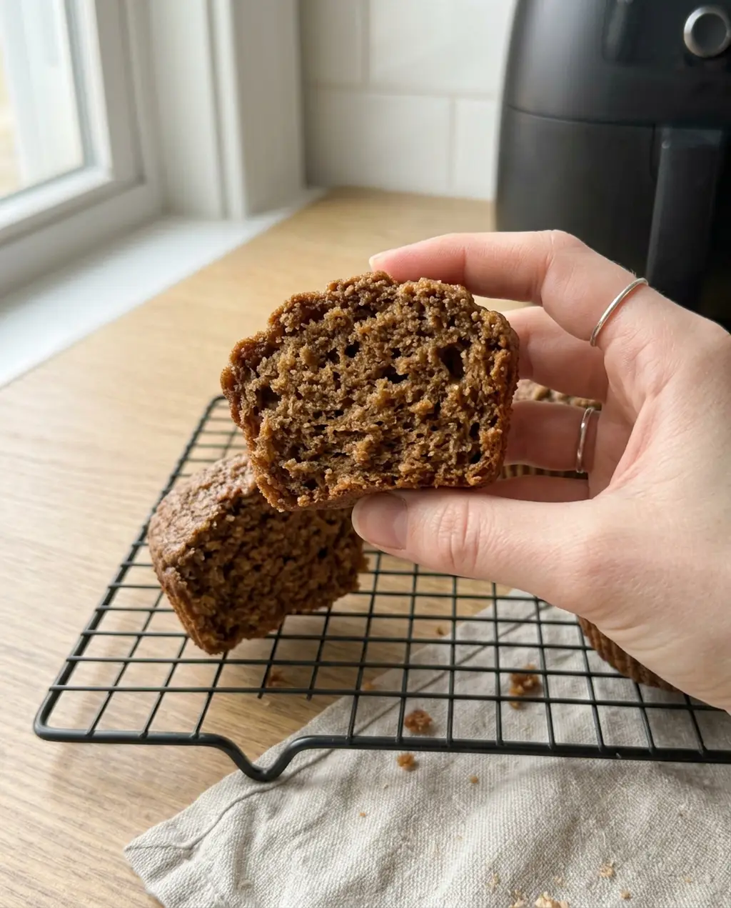 A hand holding a bran muffin split in half to show a moist, even brown crumb, over a cooling rack