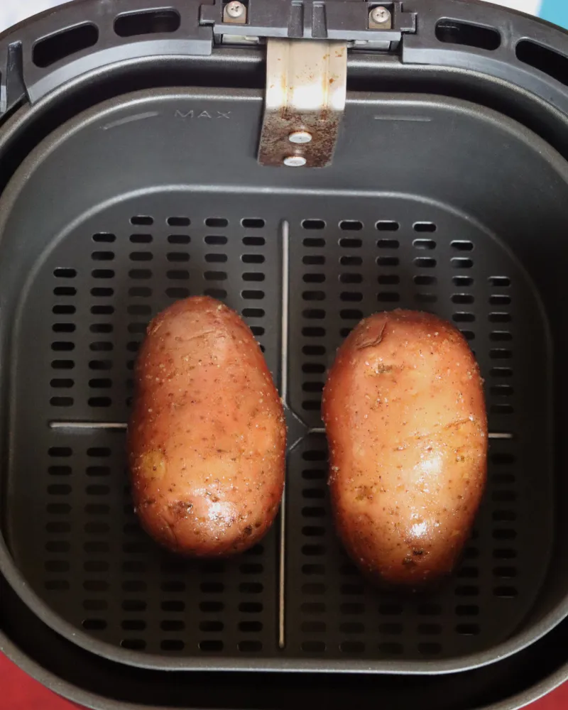 Two oiled and seasoned russet potatoes side by side in an air fryer basket before cooking
