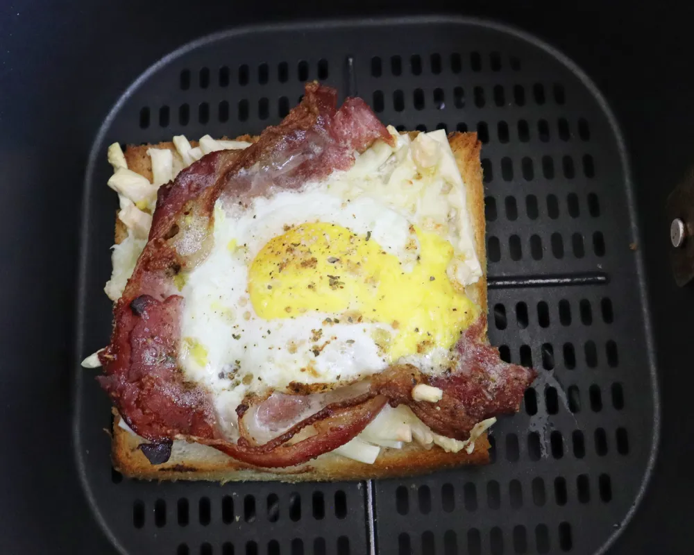 The cooked bacon and egg toast in the air fryer basket with the white set and the yolk still soft