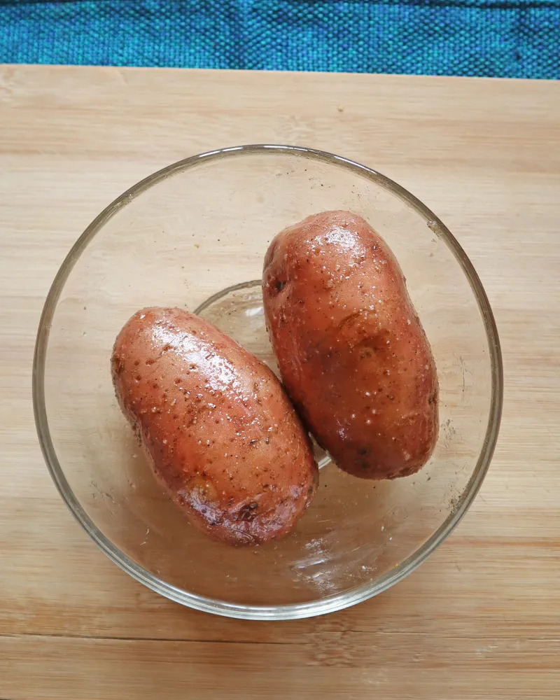 Two russet potatoes tossed in oil and seasoning in a glass bowl, skins glossy