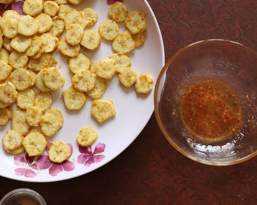 Green banana slices tossed in oil and spices spread out on a white plate, with the empty mixing bowl alongside