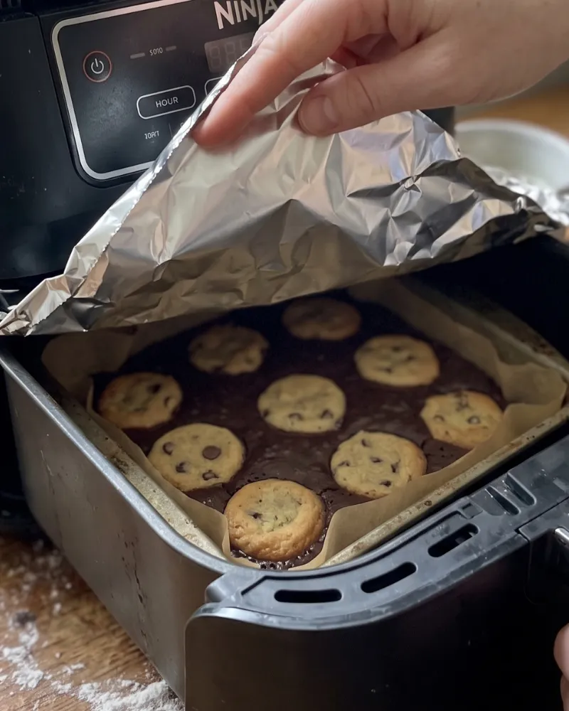 A hand lifting a sheet of foil off a pan of baking brookies inside a Ninja air fryer basket