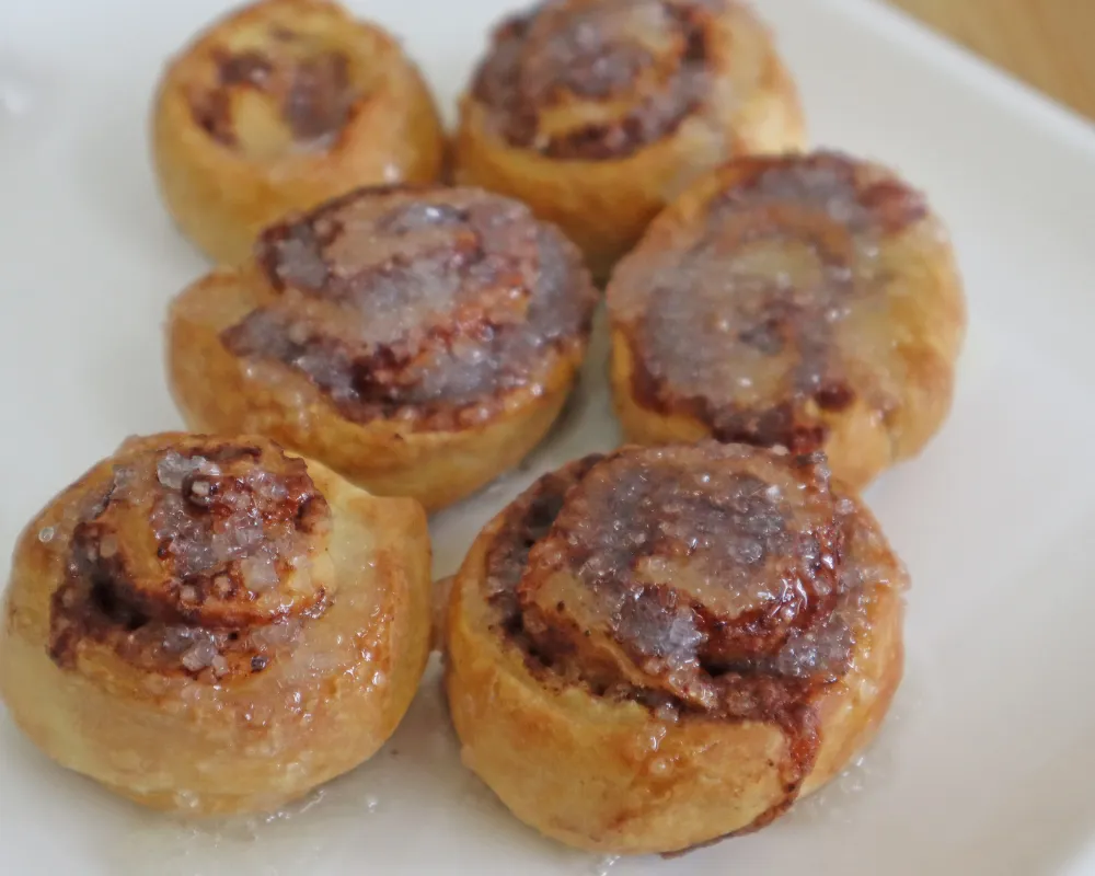 Six glazed cinnamon rolls on a white plate with a crackled sugar topping