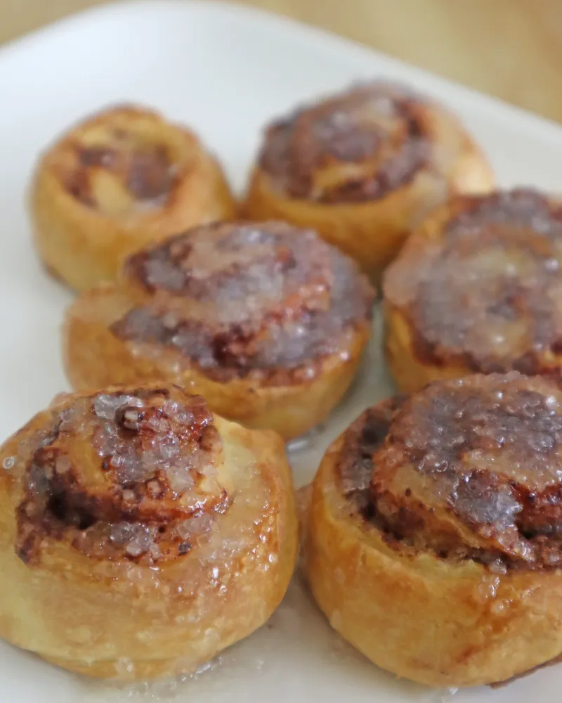 Six iced cinnamon rolls on a white plate with sugar glaze pooling on the swirls
