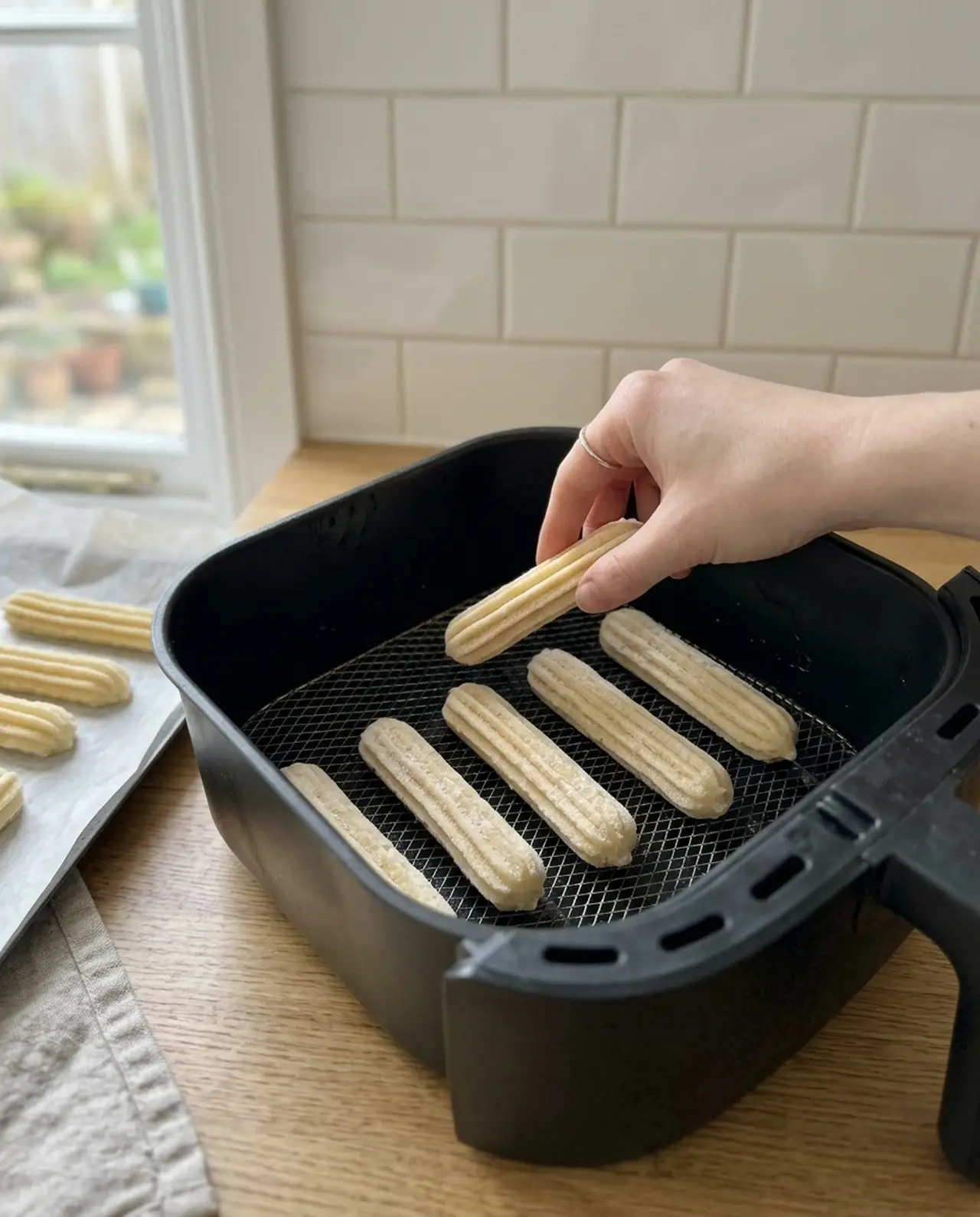 A hand placing frozen piped churro sticks in a row inside a greased air fryer basket, with more sticks waiting on a parchment tray