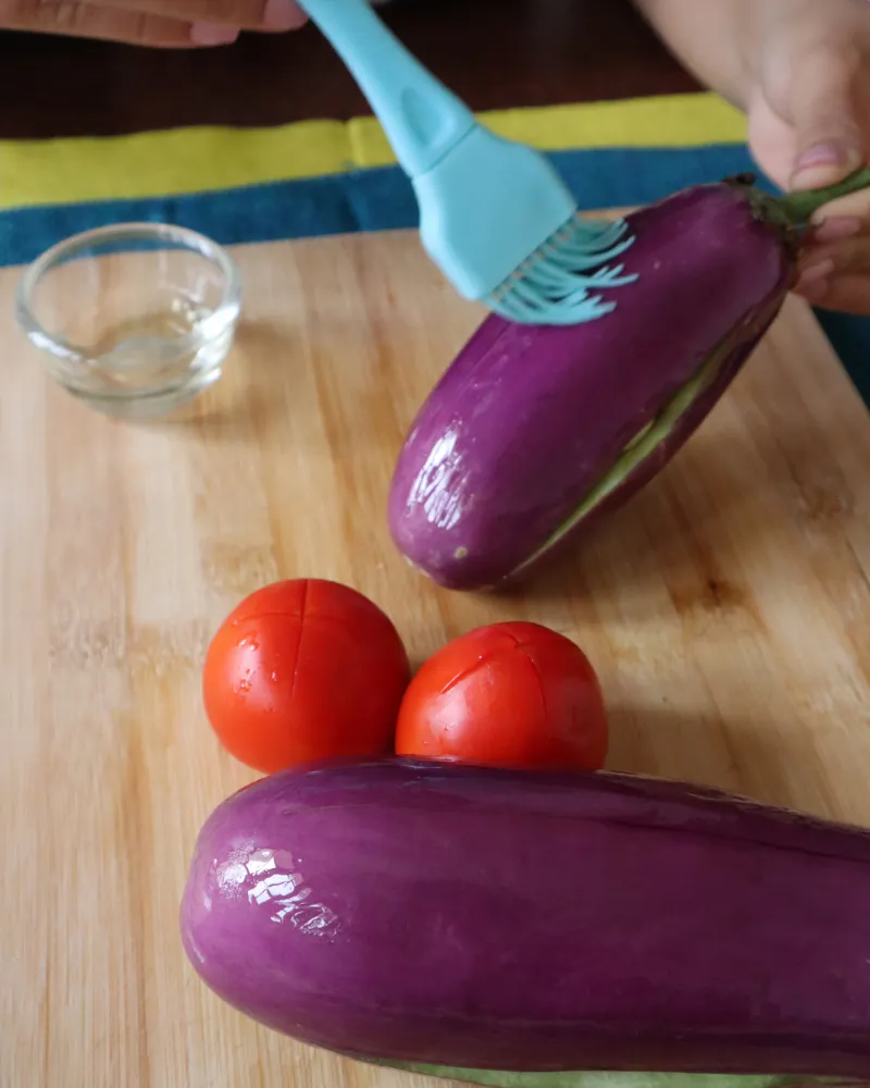 Brushing oil onto a whole eggplant on a wooden board, with two scored tomatoes alongside