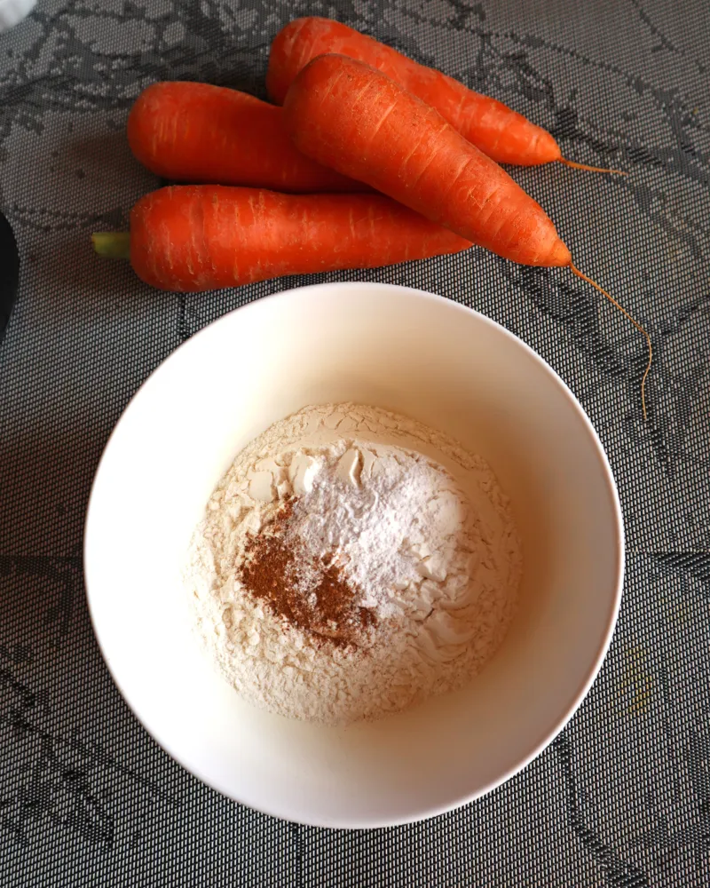 Flour, baking powder, cinnamon and salt whisked together in a white bowl beside fresh carrots