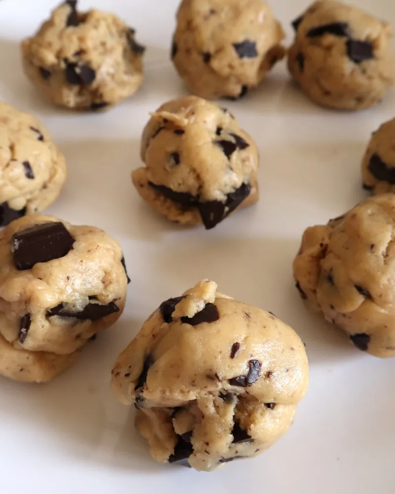 Twelve portioned balls of chocolate chip cookie dough spaced on a white plate before chilling