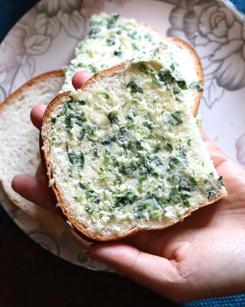 A hand holding a thick slice of sandwich bread spread edge to edge with garlic parsley butter