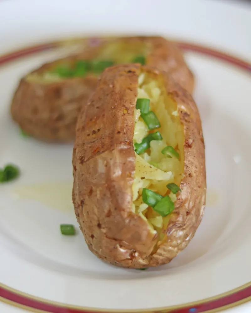 Two finished air fryer baked potatoes on a white plate, split and topped with spring onion