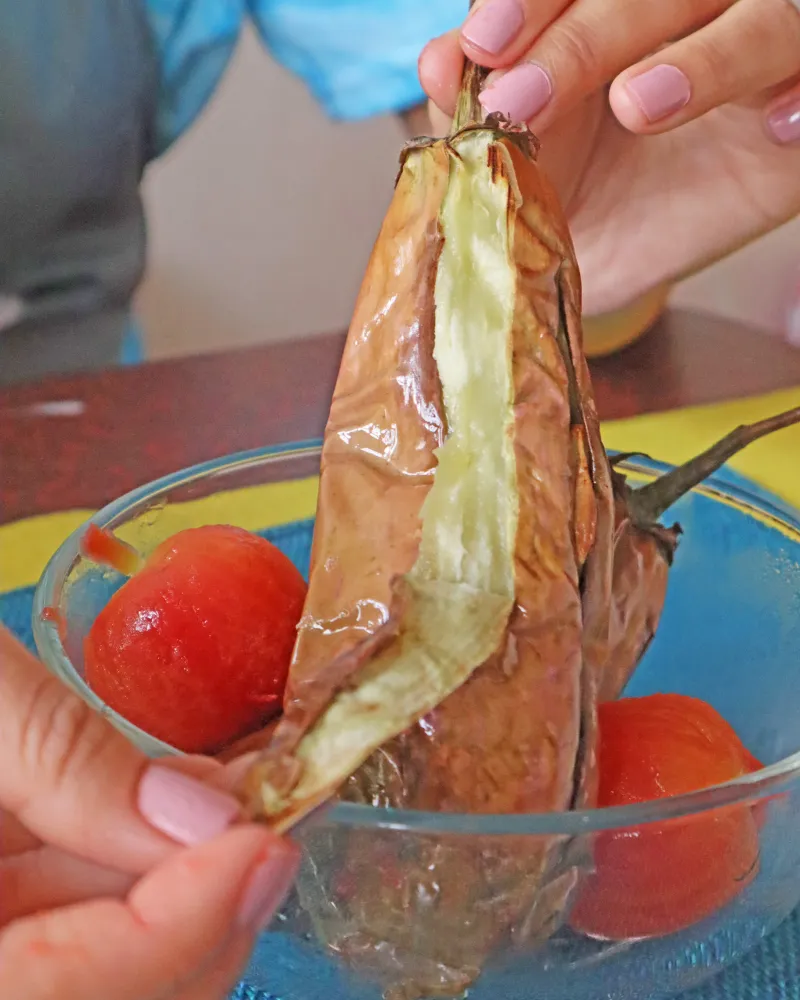 Peeling the blistered skin away from a roasted eggplant over a bowl of peeled roasted tomatoes