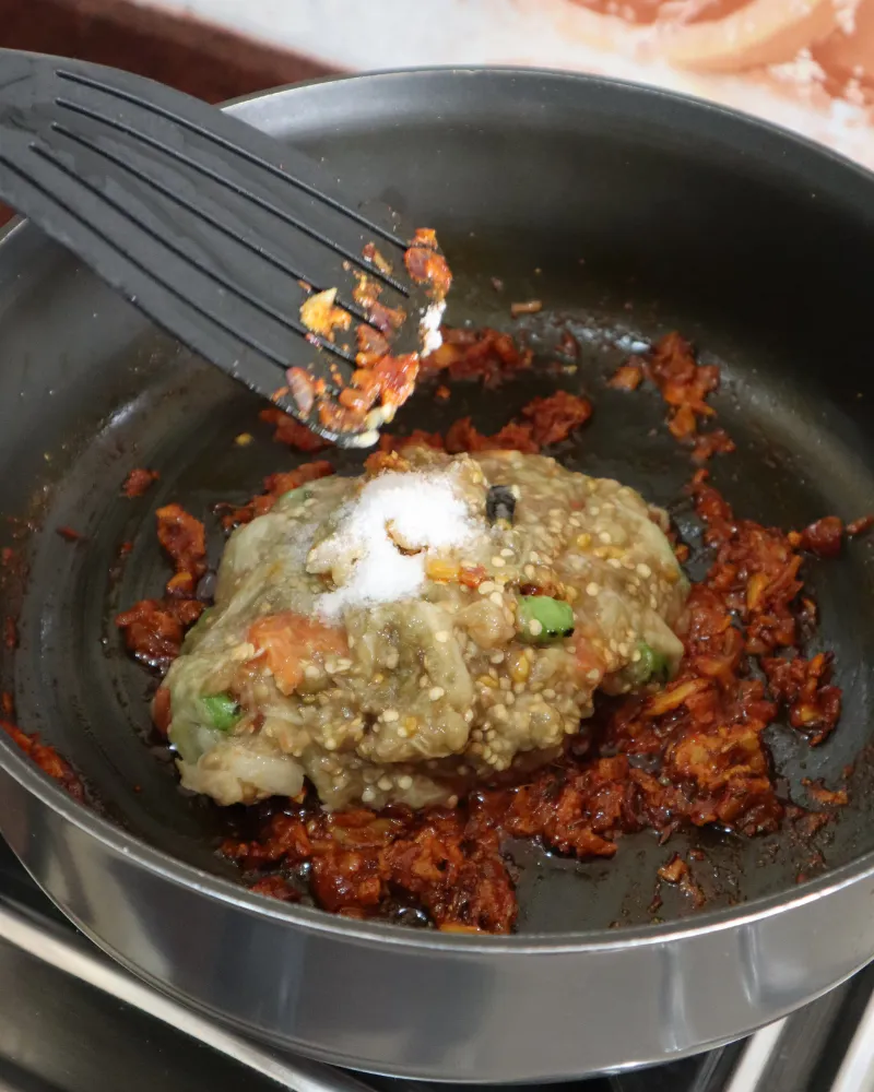 Mashed roasted eggplant being added to a pan of cooked onion and tomato masala, with salt on top